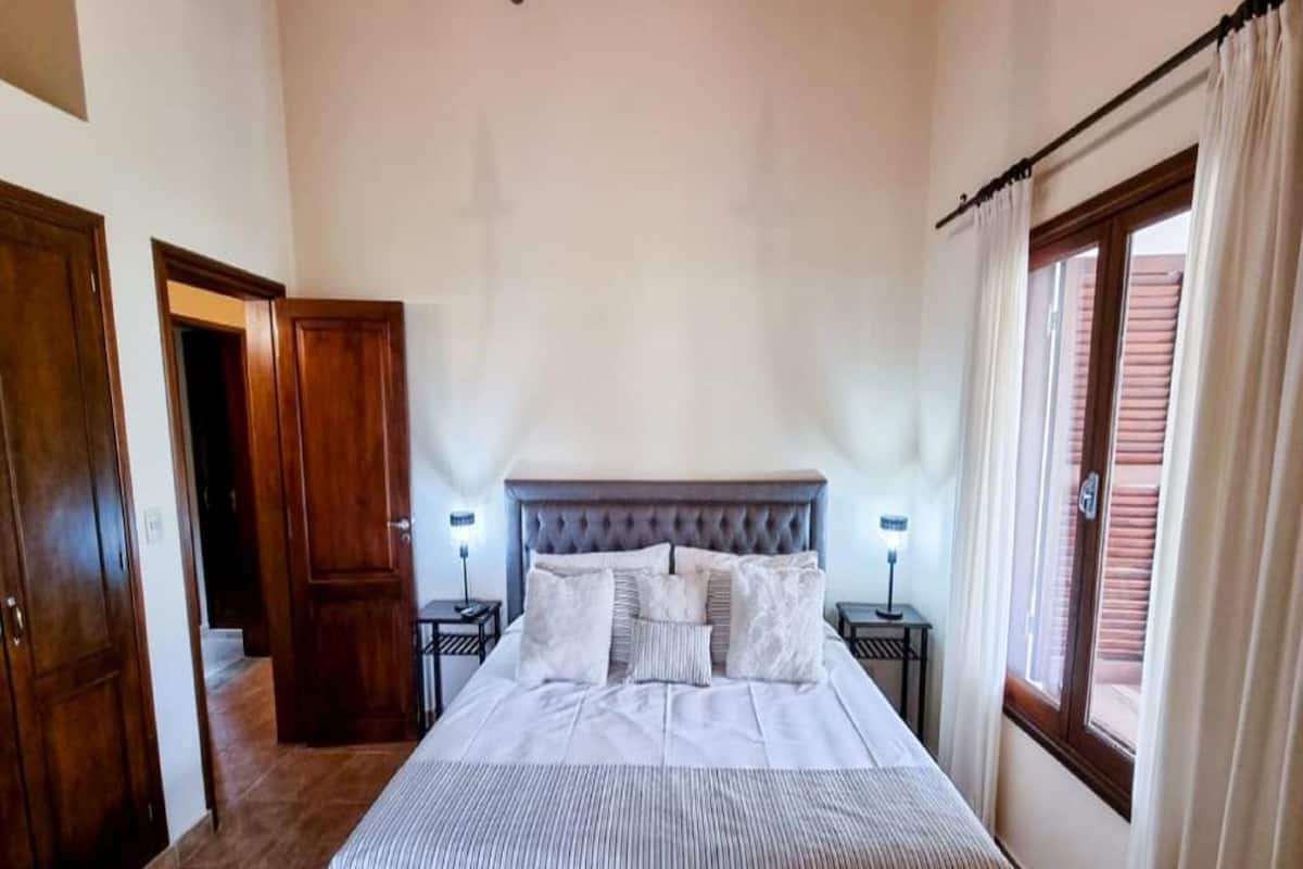 Ein Schlafzimmer mit einem großen Bett, zwei Nachttischen mit Lampen, einer Kandelaber und einem Fenster mit Vorhängen.