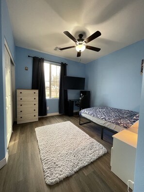 A bedroom with a bed, a desk, a dresser, a ceiling fan, and a window with curtains.