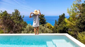 A person standing at the edge of a swimming pool overlooking a clear blue sea with a view of the ocean and surrounding greenery. - Villa Dalula in Agios Nikitas (Lefkada)