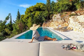 Una persona descansando junto a una piscina, con un muro de piedra y árboles al fondo.