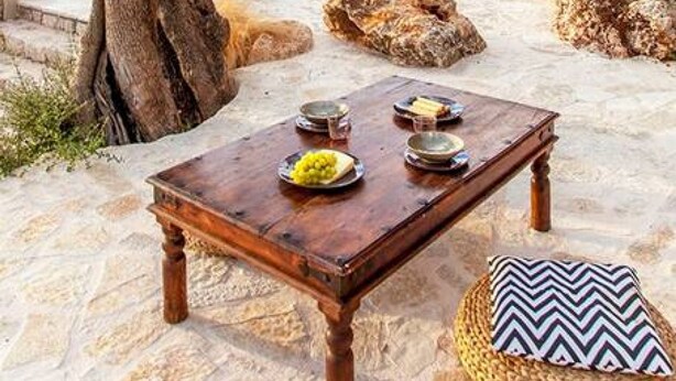 An outdoor dining area with a table set for two, surrounded by natural stone flooring and a view of a pool and greenery. - Villa Dalula in Agios Nikitas (Lefkada)