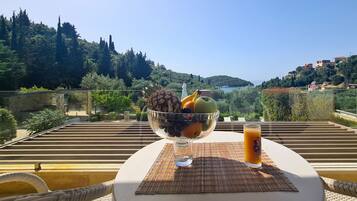 A table with a fruit bowl and a glass of orange juice on a balcony overlooking a scenic view of hills and trees.