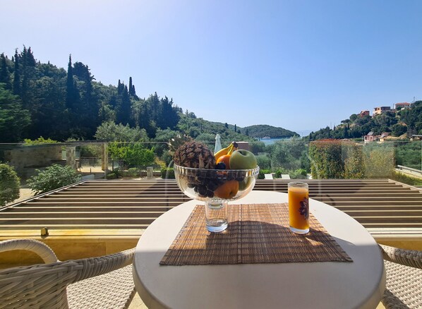 Una mesa con un frutero y un vaso de jugo de naranja en un balcón con vistas panorámicas a colinas y árboles.
