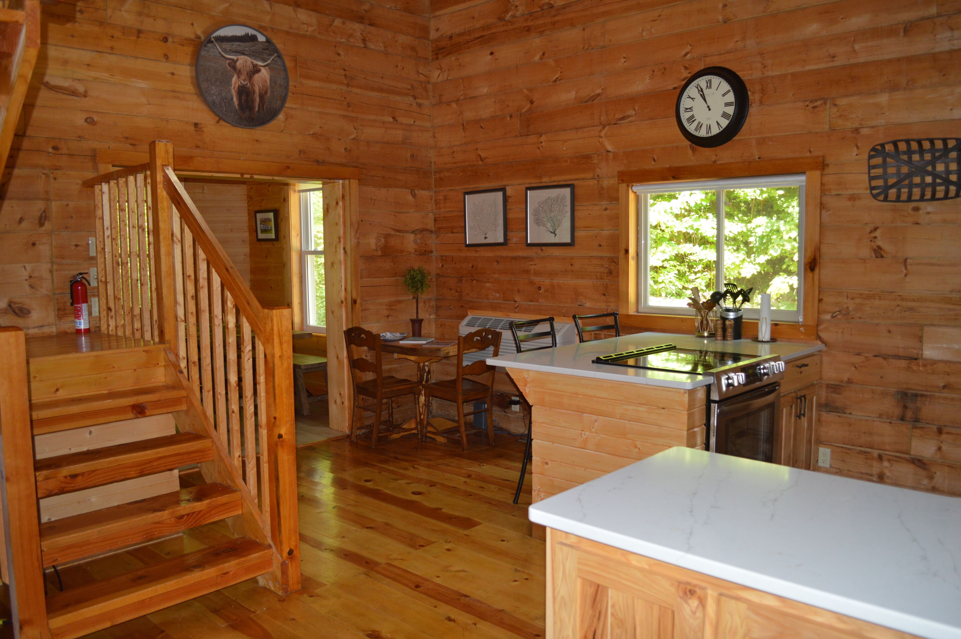 A wooden cabin interior with a kitchen area, dining table, chairs, a staircase, and a window with a view of trees.
