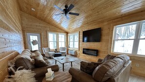 A cozy log cabin interior with a wooden ceiling, a ceiling fan, a large TV mounted on the wall, and a fireplace. - Cozy Secluded Cabin w/ Hot Tub, Relax, Unwind & Stargaze (Ferryville)