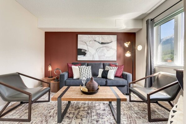 Una sala de estar moderna con un sofá gris, dos sillones, una mesa de centro de madera y un cuadro abstracto grande en la pared.