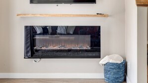 A modern living room with a flat-screen TV mounted above a fireplace, a wooden shelf below, and a blue woven basket on the floor.
