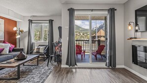 A modern living room with a sofa, chairs, a coffee table, and a fireplace. There is a sliding glass door leading to a balcony with red chairs.