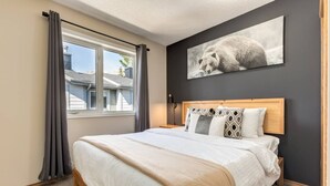 A bedroom with a large bed, a window with curtains, a headboard, and a wall-mounted picture of a bear.