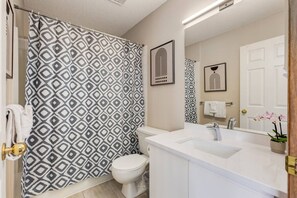 Un baño con cortina de ducha estampada, inodoro, lavamanos con espejo y puerta con una toalla colgada.