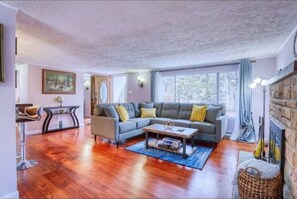 Amplia sala de estar con piso de madera, un sofá seccional gris con cojines amarillos, una mesa de centro y un ventanal con cortinas.