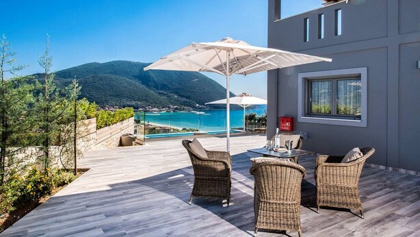 Patio esterno moderno con mobili in vimini, un tavolo e un grande ombrellone. Si affaccia su una zona costiera panoramica con montagne e un mare blu cristallino.