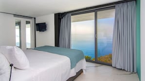 A bedroom with a bed, a TV mounted on the wall, and a large sliding glass door leading to a balcony with a view of the sea. - Villa Blue Ionian - Syvota (Igoumenitsa)