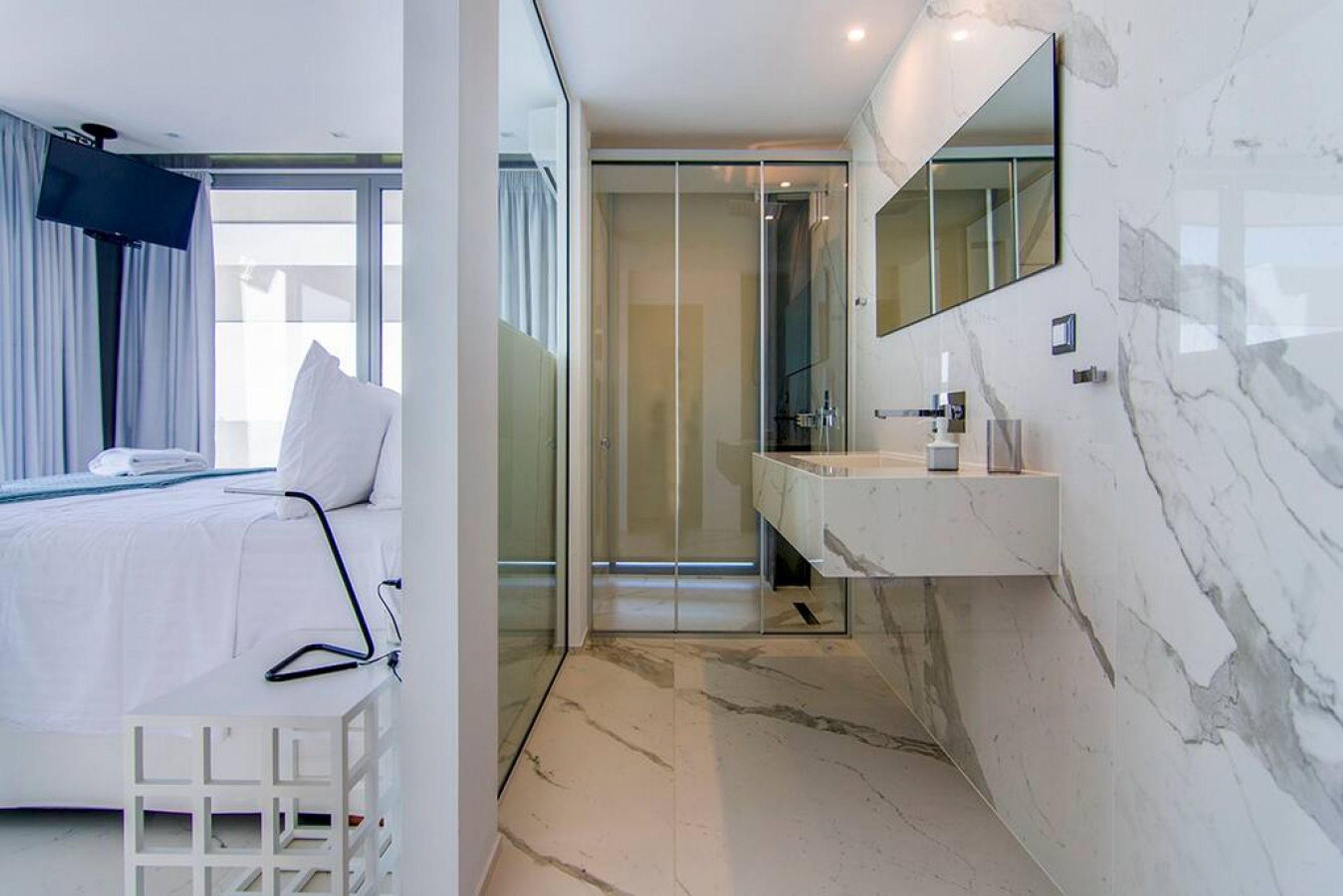 A modern hotel room with a bed, a large mirror, a bathroom with a marble countertop, and a glass shower enclosure.