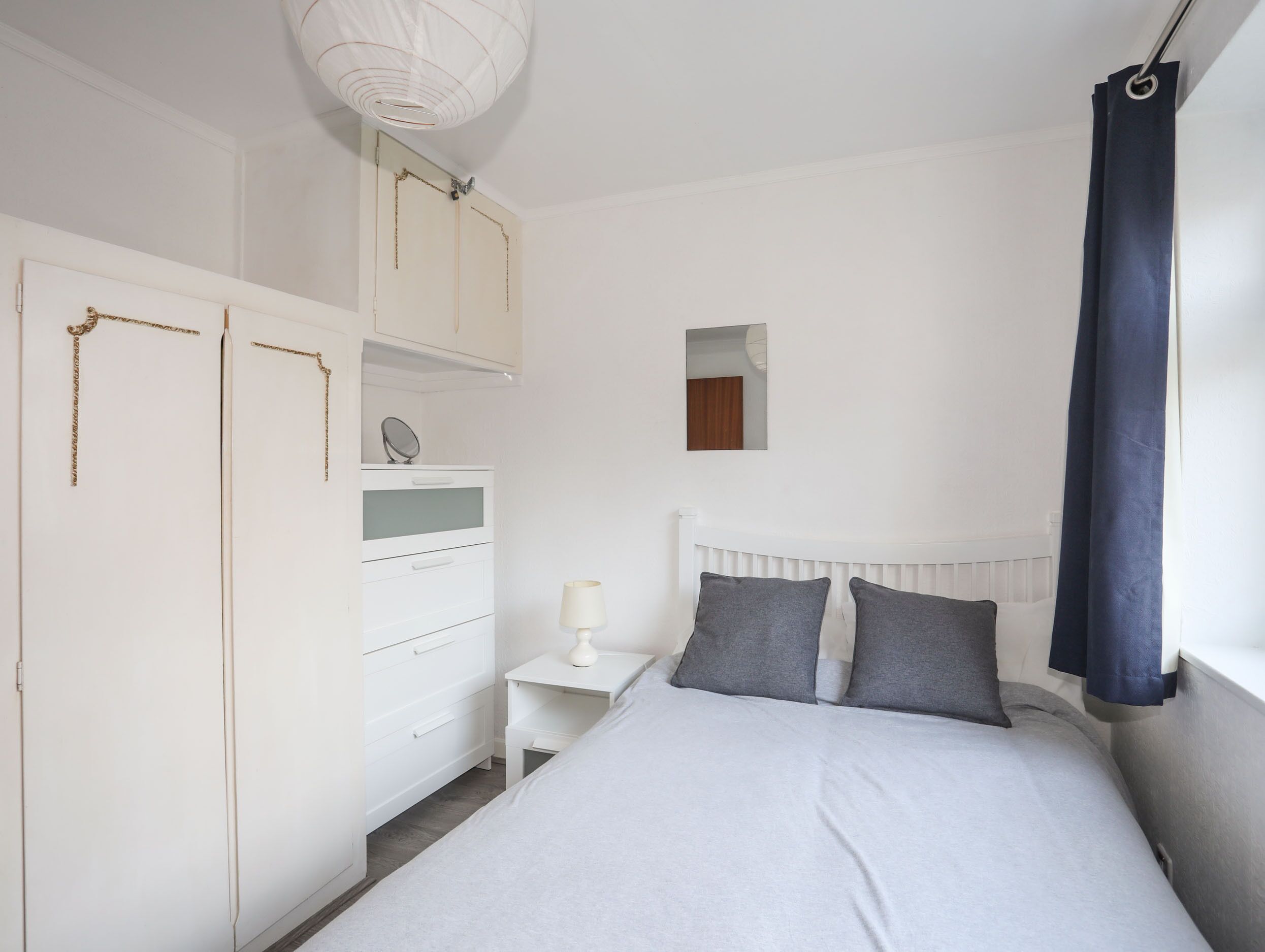 Quarto com cama, mesa de cabeceira, penteadeira, espelho e janela com cortinas.