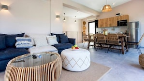 Ein modernes Wohnzimmer mit einer Couch, zwei Hocker und einem Glastisch. Ein Essbereich mit Korbstühlen und eine Küche mit Holdekor-Schränken und Edelstahlgeräten sind ebenfalls zu sehen.
