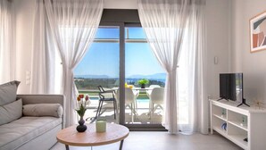 A modern living room with a sofa, a round table with a vase, a TV on a white stand, and a balcony with a view of the landscape. - Villa Perseids Lumiere (Preveza)