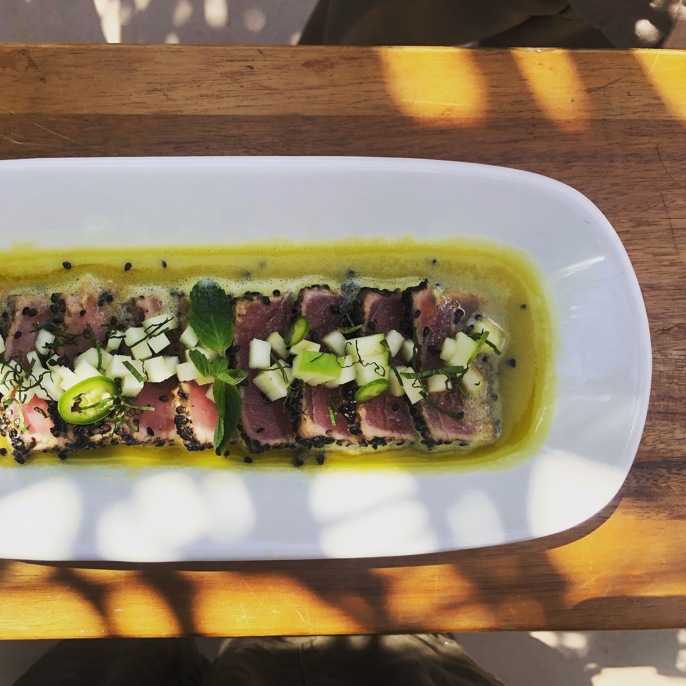 Un plato blanco rectangular con un guiso de carne en rodajas, verduras picadas y hierbas, bañado en salsa amarilla, sobre una mesa de madera.