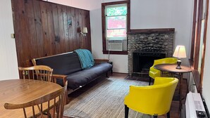 A cozy living room with a wooden wall, a sofa, a fireplace, a table, and two yellow chairs. - Waterfront Pine Cabin near Saratoga, Lake George (Corinth)