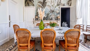 Ein Esszimmer mit einem weißen Tisch, gedeckt für sechs Personen, umgeben von Korbstühlen. Hinter dem Tisch befindet sich ein großes tropisches Wandbild.