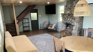 Un salon chaleureux comprenant un canapé, une table à manger avec des chaises, une cheminée en pierre, un escalier et un téléviseur fixé au mur.
