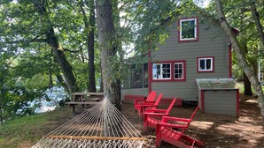 Une cabane avec un hamac, des chaises Adirondack rouges et une table de pique-nique, le tout situé près d’un lac.