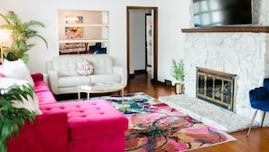 A living room with a white sofa, a pink ottoman, a fireplace, and a TV mounted above it.