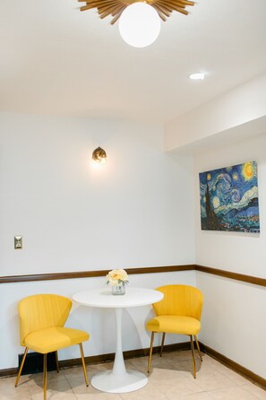 Una habitación con una mesa redonda blanca, dos sillas amarillas, una lámpara fijada a la pared y un cuadro de un cielo nocturno.