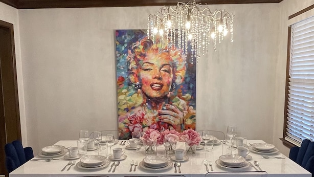 A dining room with a long table set for six, clear chairs, a chandelier, and a large painting on the wall.