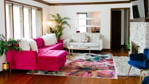 A living room with a pink sofa, a white armchair, and a blue armchair. There is a bookshelf and a fireplace.