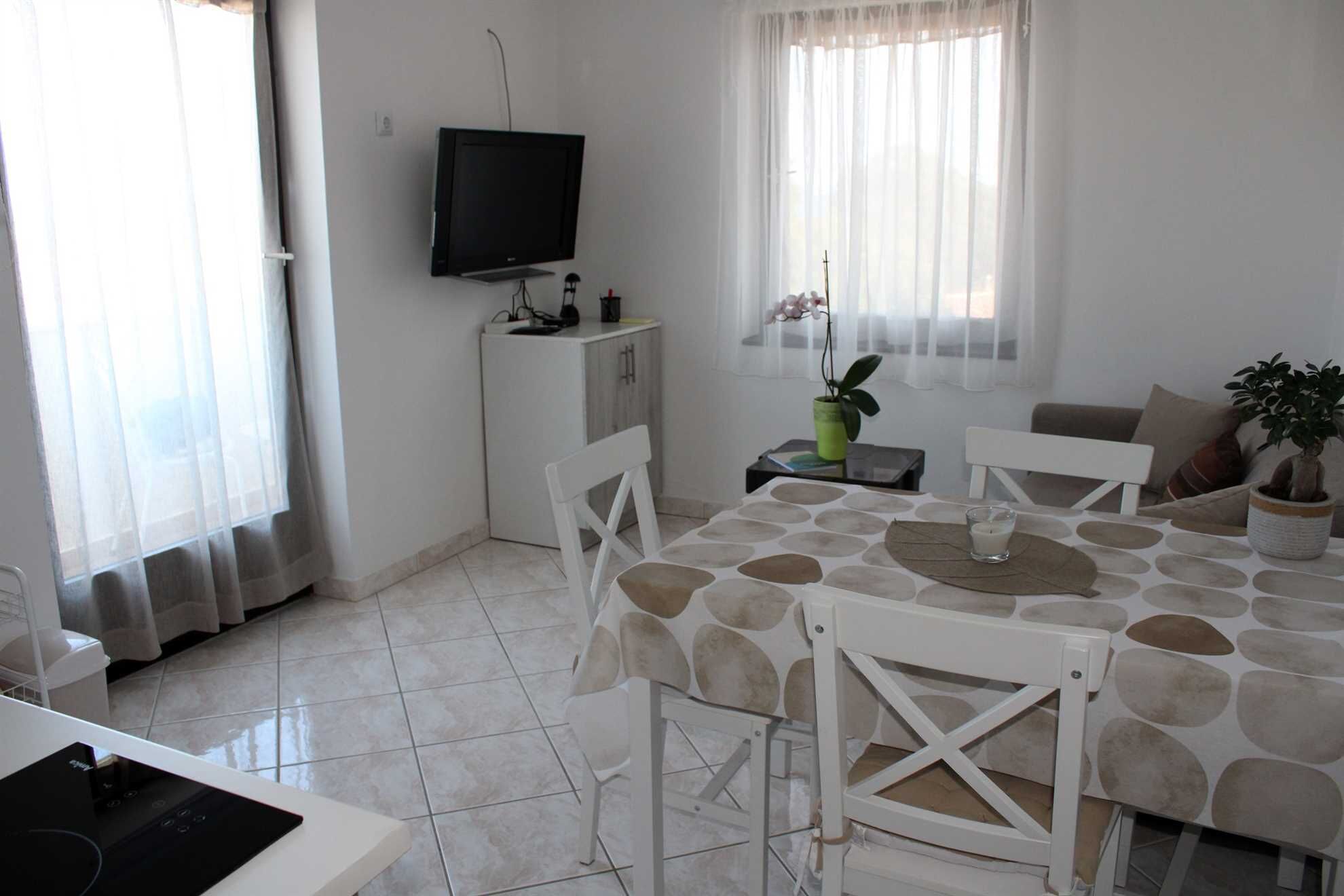 A dining area with a table set for four, a television on a stand, a small cabinet, and a potted plant.