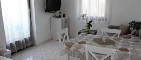 A dining area with a table set for four, a television on a stand, a small cabinet, and a potted plant.