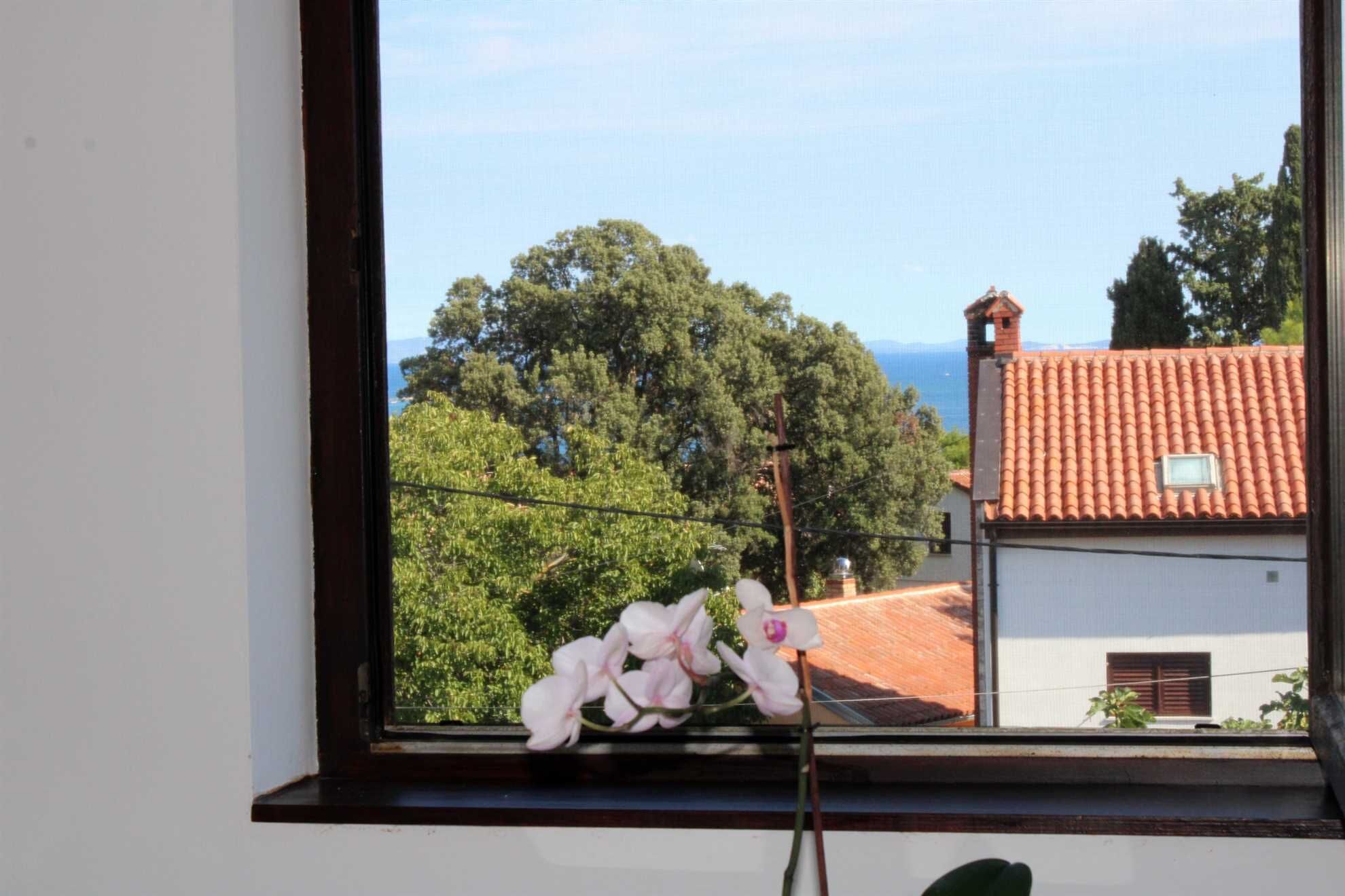 A window with a view of a building with a red-tiled roof and a balcony with a plant.