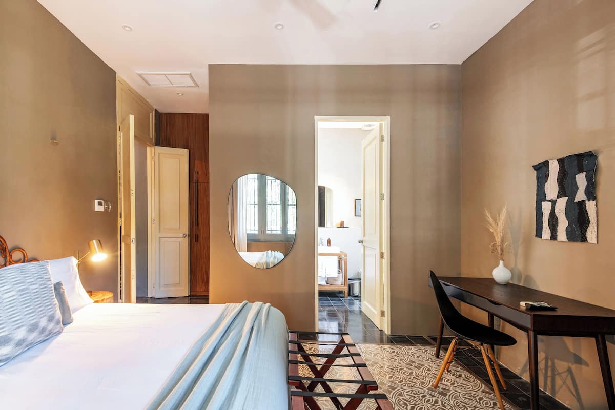 A modern hotel room with a bed, desk, and chair, a mirror, and a view into the bathroom.