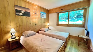 Une pièce aux murs lambrissés, avec deux lits, une fenêtre donnant sur les arbres et une lampe sur une table de chevet.