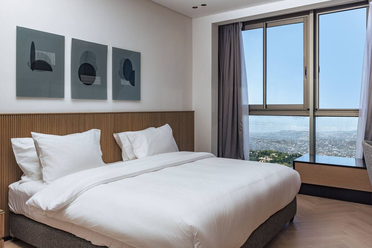 Una habitación de hotel moderna con una cama grande, un cabecero de madera y vistas a la ciudad a través de ventanas de techo a suelo.