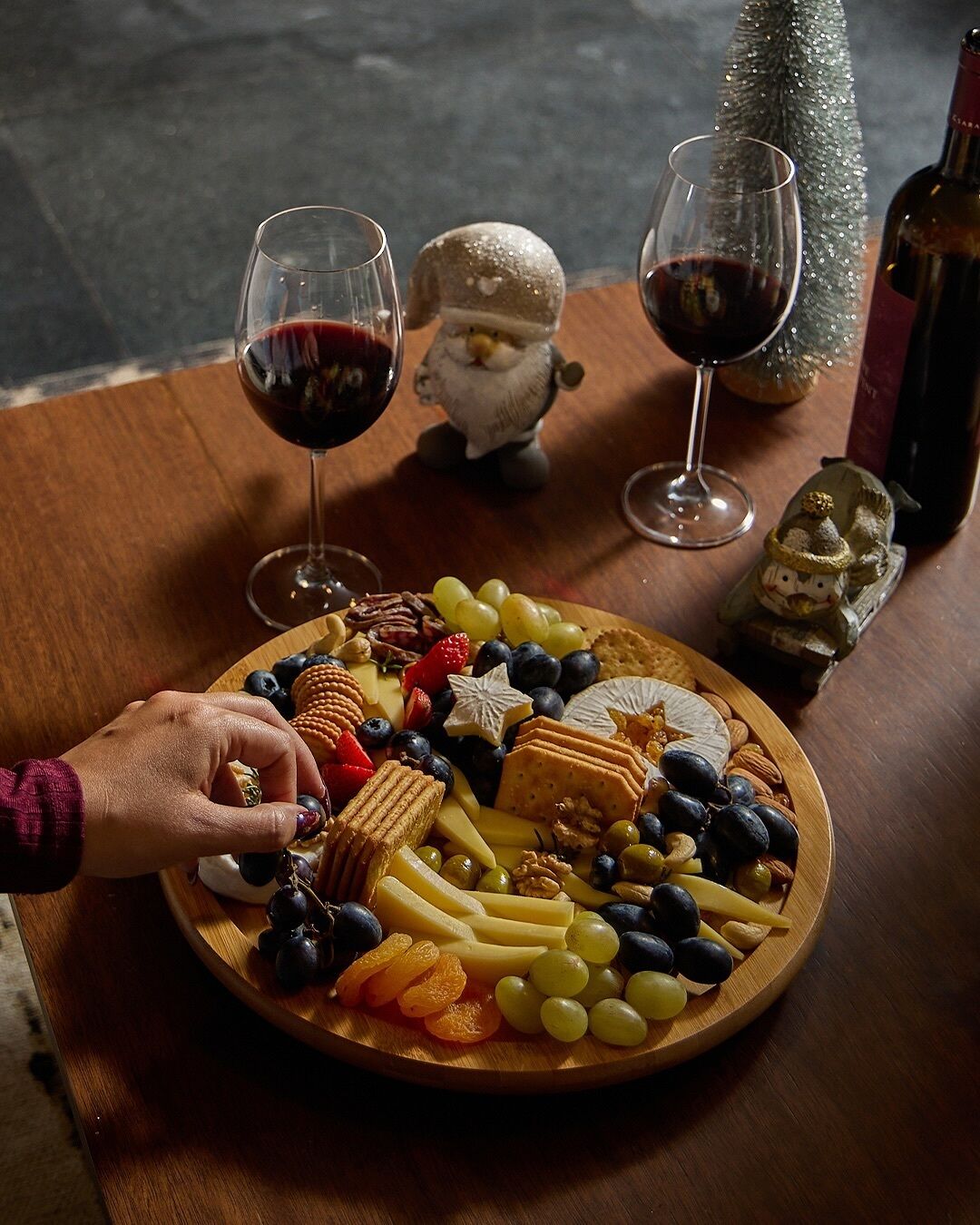Una tabla de madera con una variedad de quesos, frutas y frutos secos. Hay dos copas de vino tinto sobre la mesa, y se ve una pequeña decoración navideña.