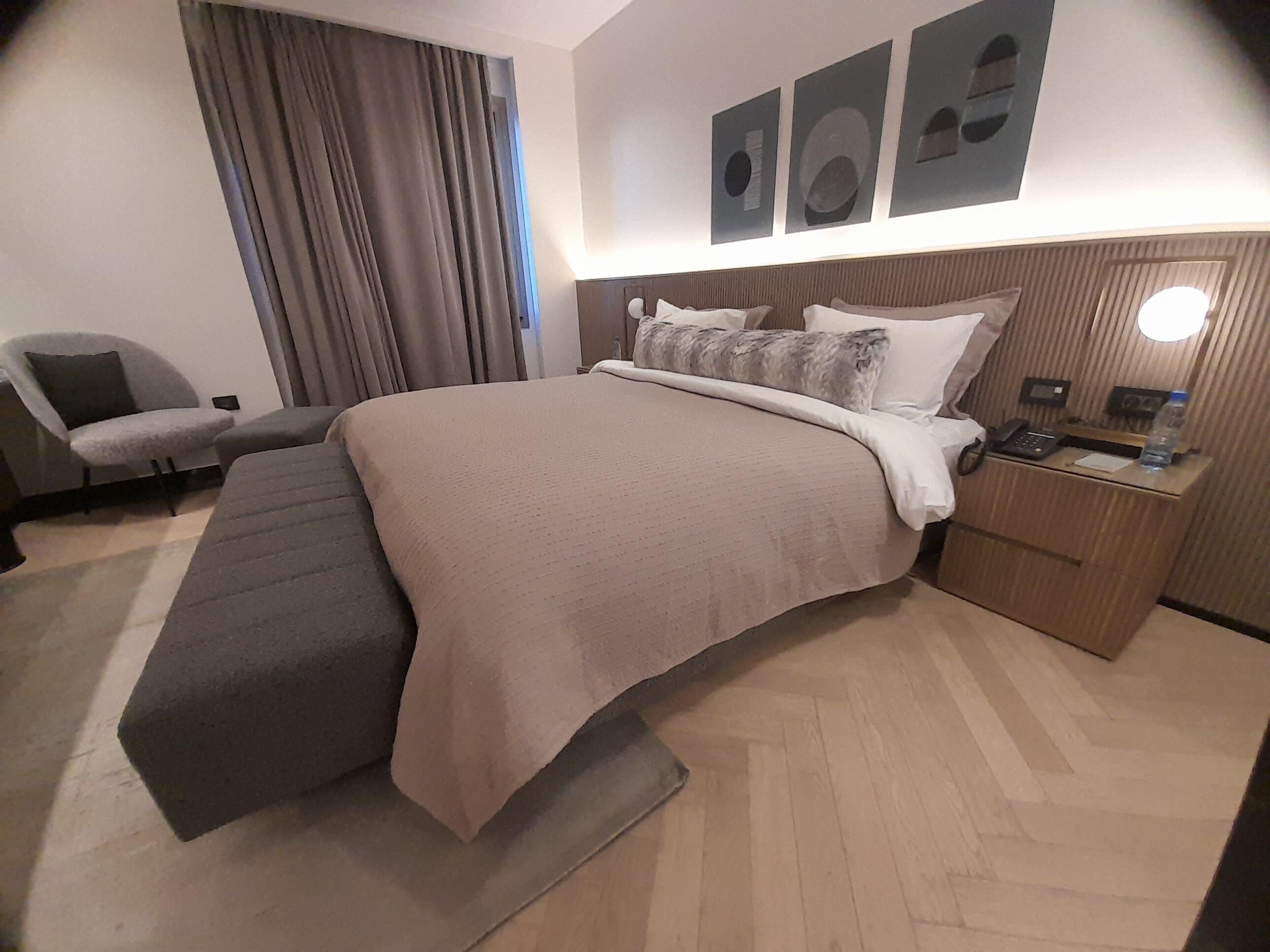 Et moderne hotelværelse med en stor seng, et natbord med telefon og flaske, en lænestol og et stort vindue med gardiner.