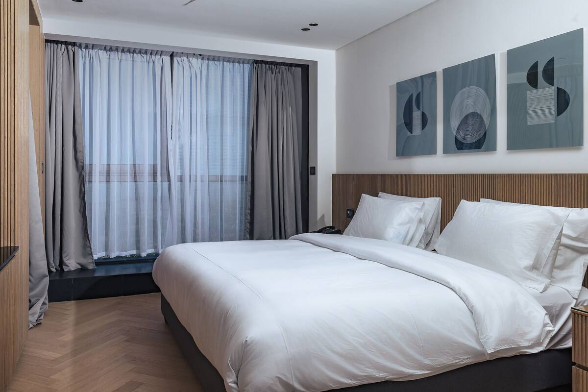 Habitación de hotel moderna con una cama grande, cabecera de madera y vistas a través de cortinas translúcidas.