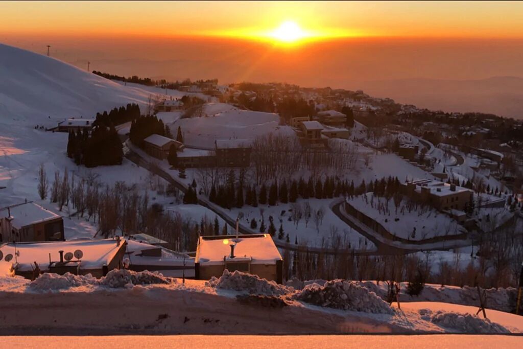 Un village enneigé, avec ses bâtiments et ses maisons, un ciel dégagé et le soleil se couchant à l’horizon.