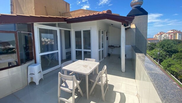 A covered outdoor patio with a table and chairs, large windows, and a view of the cityscape. - Cobertura Vista Mar Jurerê Internacional (Florianópolis)