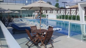 Un área de piscina en la azotea con mesa y sillas, una tumbona y barandilla de cristal.