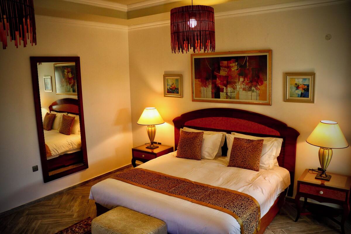 Quarto de hotel com uma cama grande, duas mesas de cabeceira com luminárias, um espelho e obras de arte nas paredes.