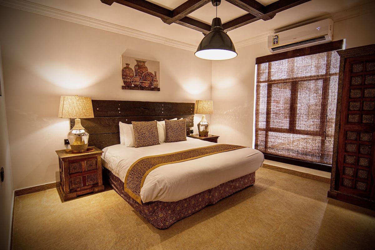 Uma cama bem arrumada com colcha estampada, duas mesas de cabeceira com abajures, uma obra de arte na parede e uma janela grande com vista.