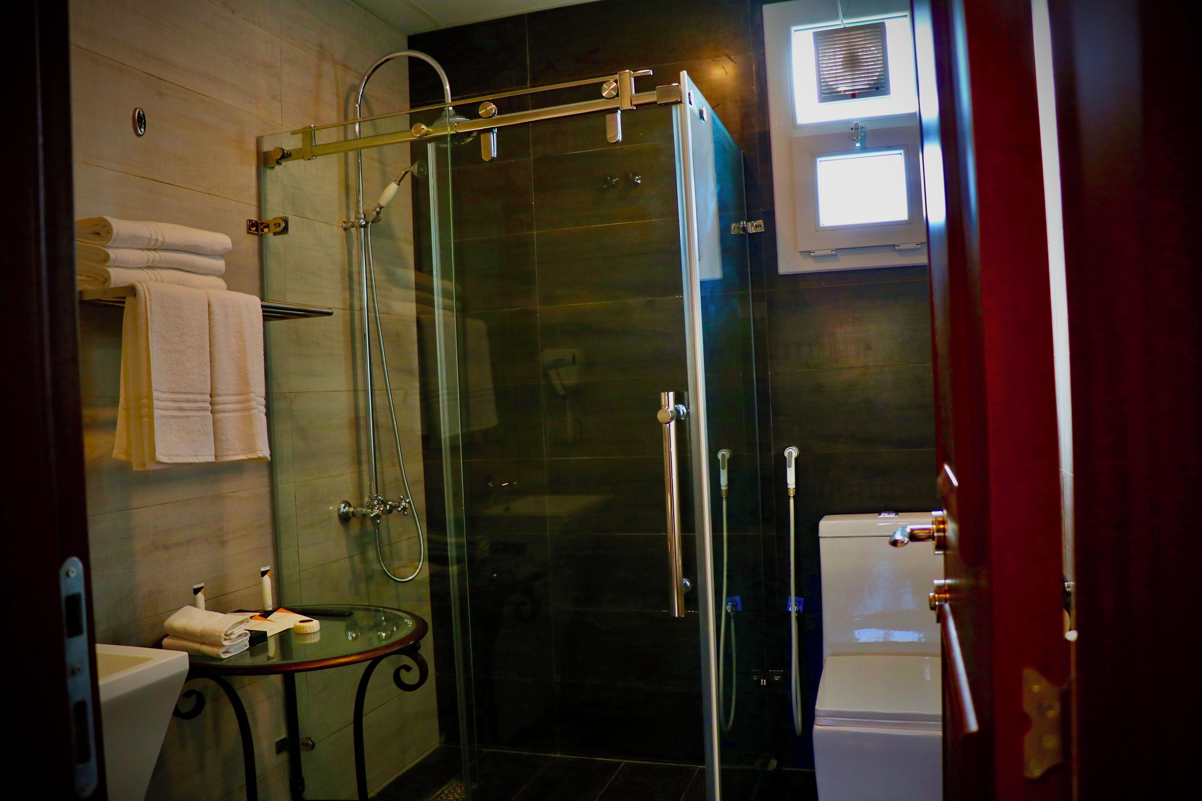 A glass-enclosed shower with a handheld showerhead, a white toilet, and a small round table with a towel and a bottle.