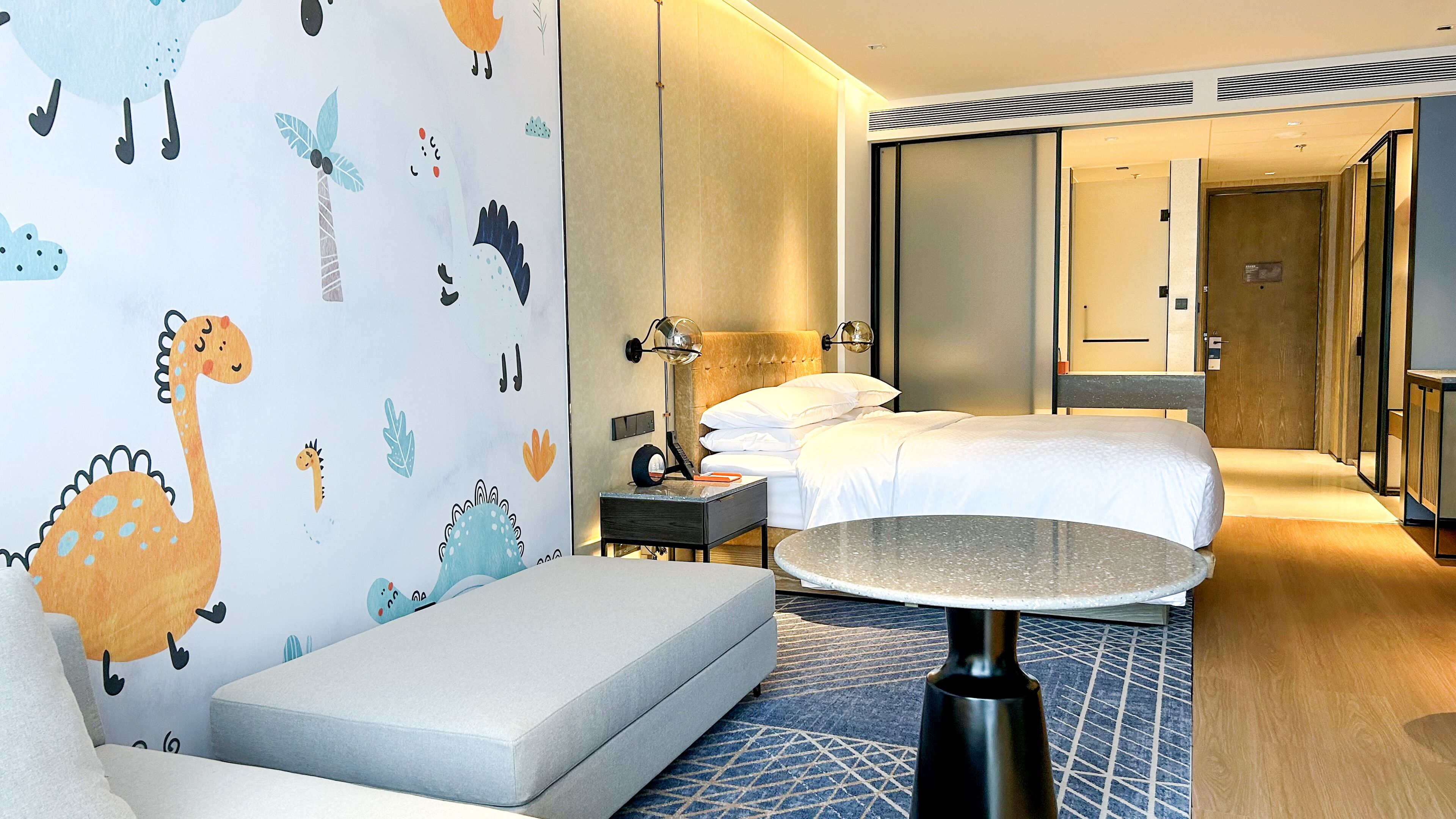 Una habitación de hotel con una cama, una mesita de noche, una lámpara, un sofá y un papel pintado con motivos de dinosaurios.