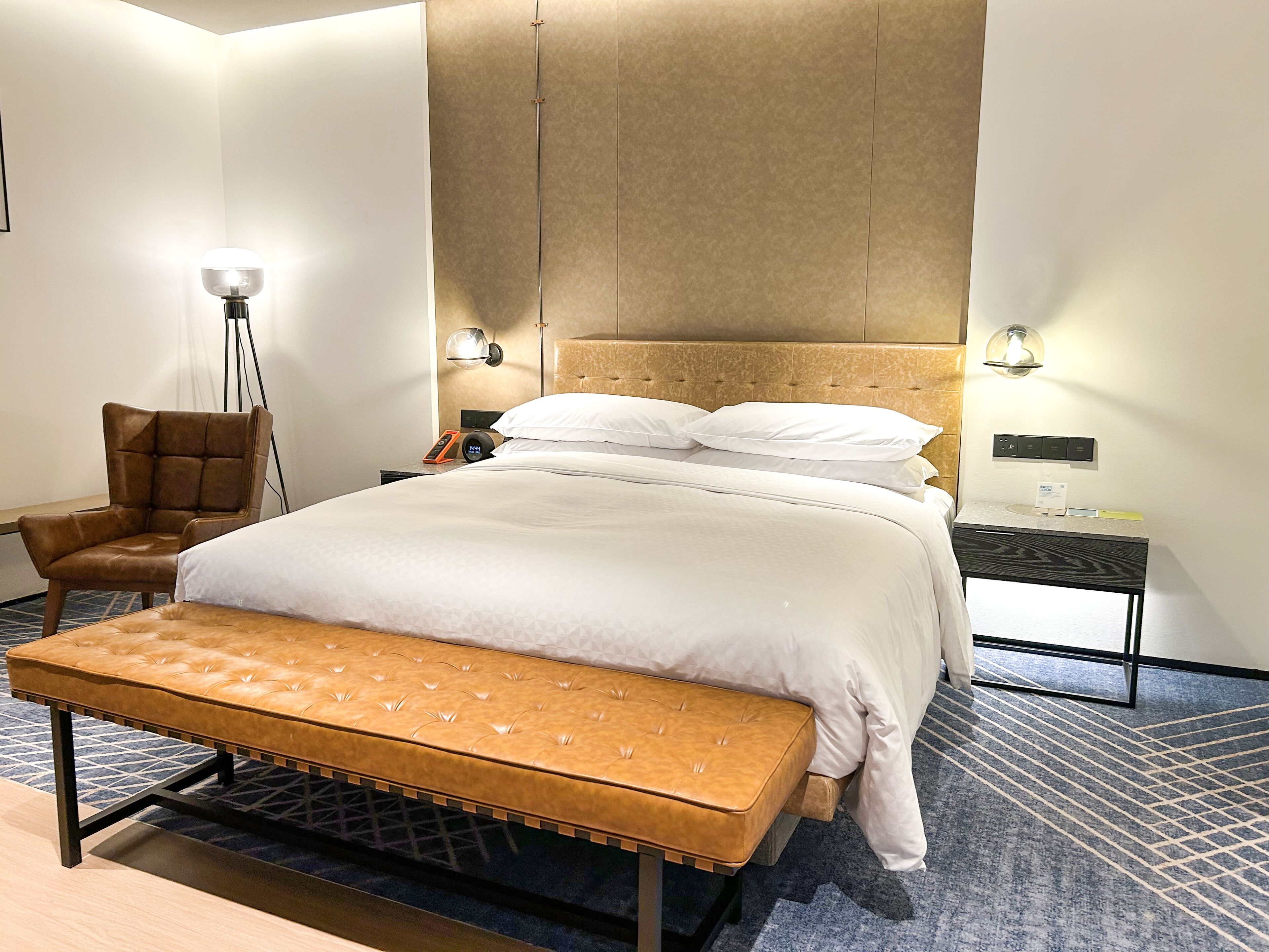 Una habitación de hotel moderna con una cama grande, un banco, una silla, lámparas de noche y una mesita de noche con un teléfono y estación de carga.