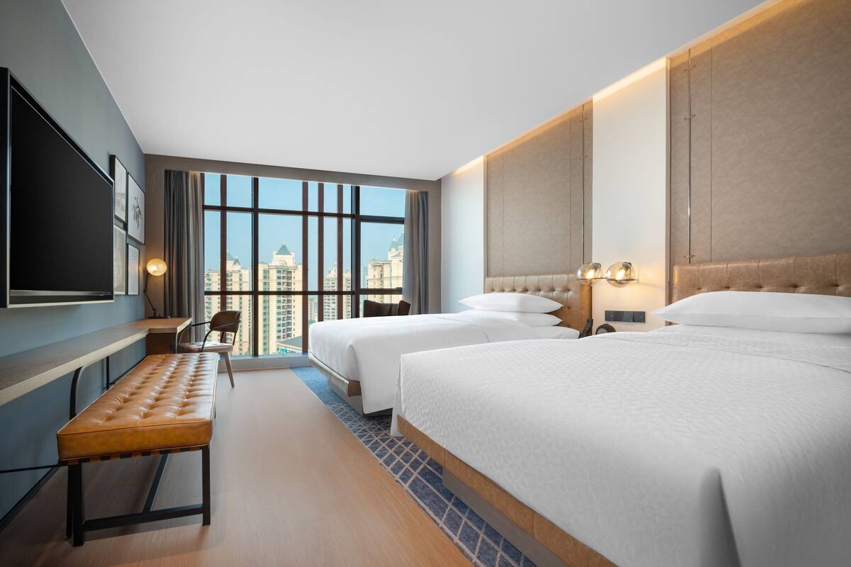 Ein Hotelzimmer mit zwei Betten, einer Bank, einem Fernseher und großen Fenstern mit Blick auf die Stadt.