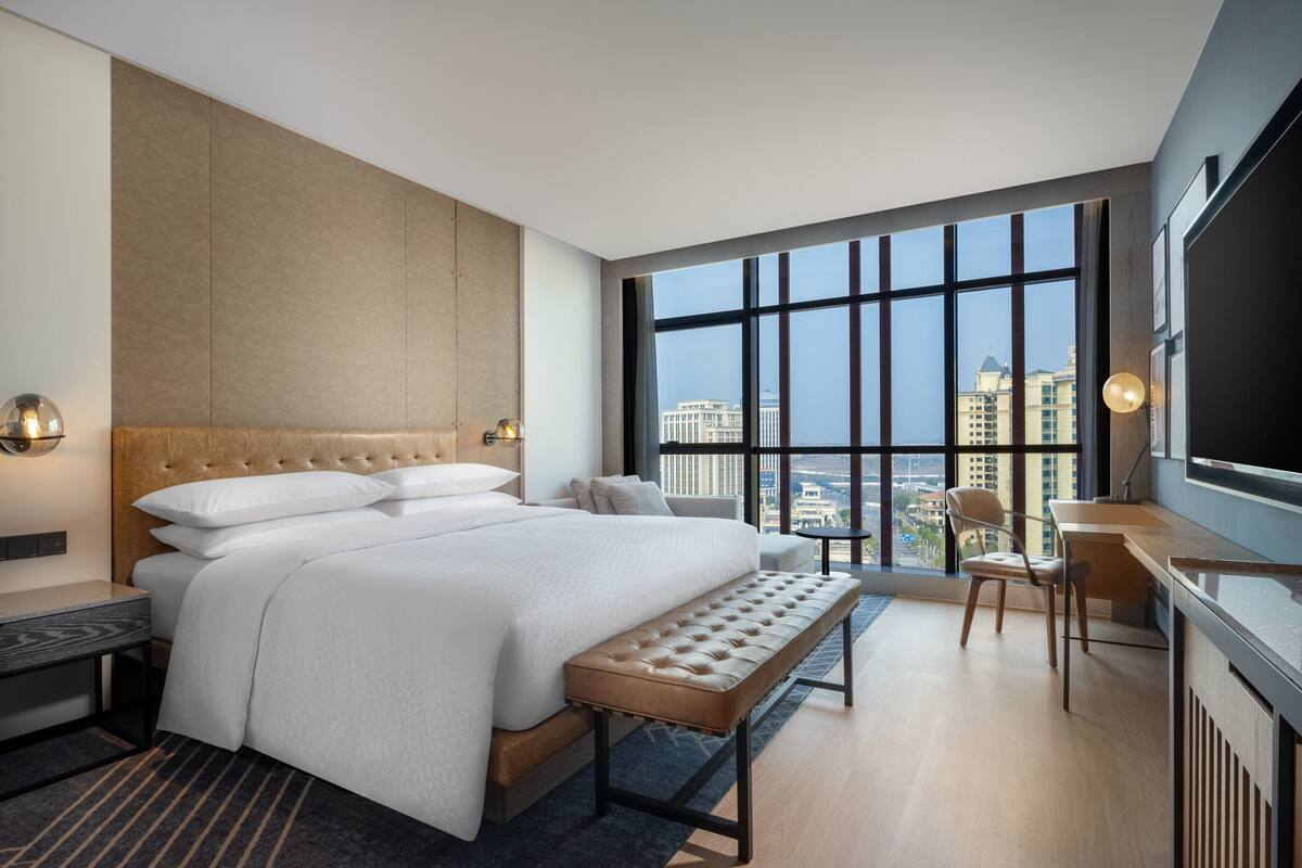 Ein modernes Hotelzimmer mit einem großen Bett, einem Schreibtisch und Blick auf die Stadt durch große Fenster.