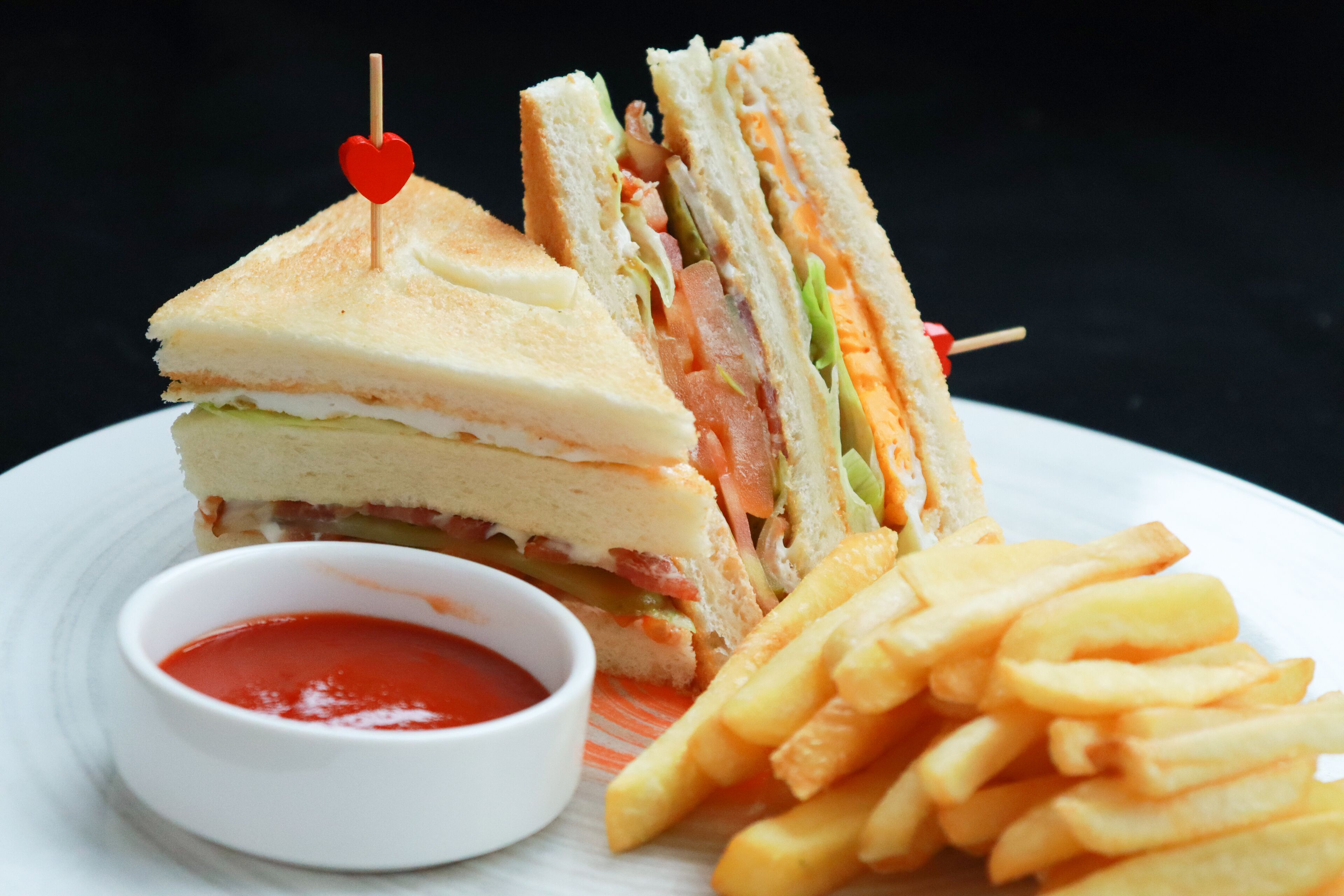 Un sandwich décoré avec un cure-dent en forme de cœur, accompagné d’un petit bol de ketchup et d’une portion de frites.